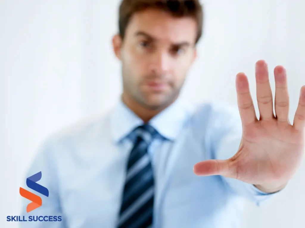 A man in a blue shirt raises his hand, signaling or greeting, with a neutral background