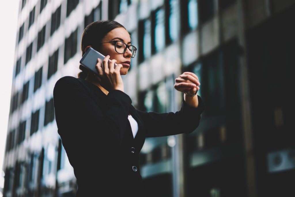 confident-businesswoman-dressed-in-elegant-apparel-looking-at-smartwatch-while-talking-on-smartphone-financial-expert-managing-time-on-wristwatch-during-mobile-conversation-promotional-background