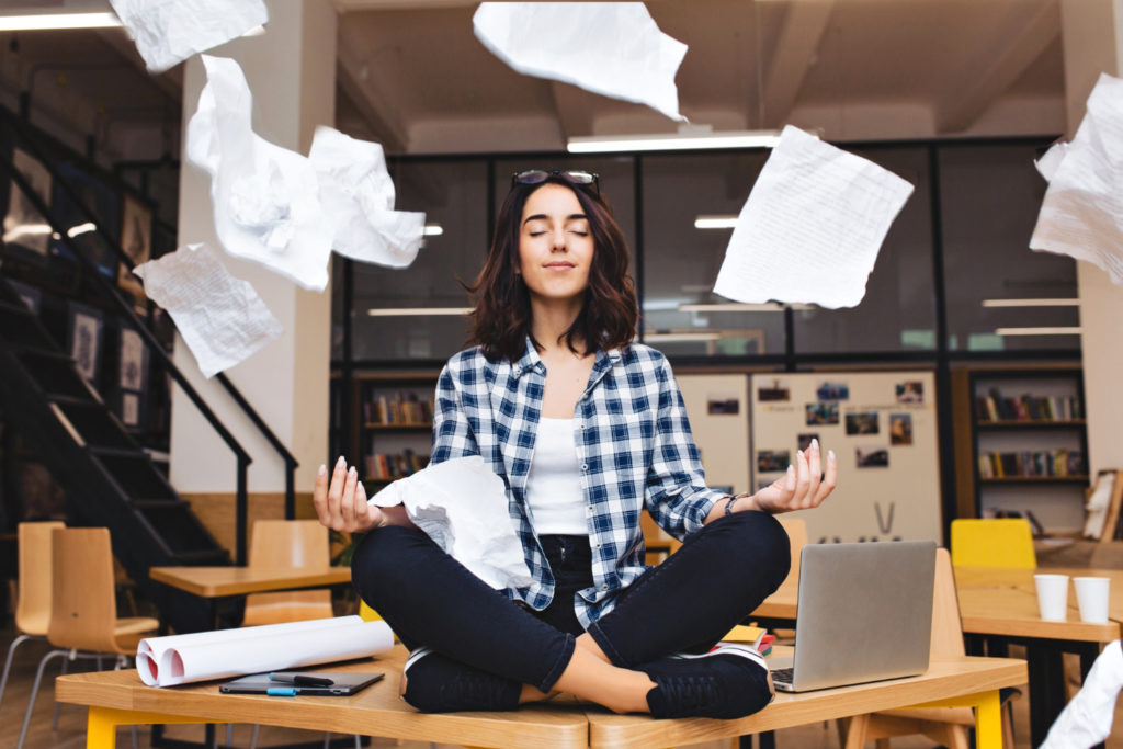 Young pretty joyful brunette woman meditating on table surround work stuff and flying papers