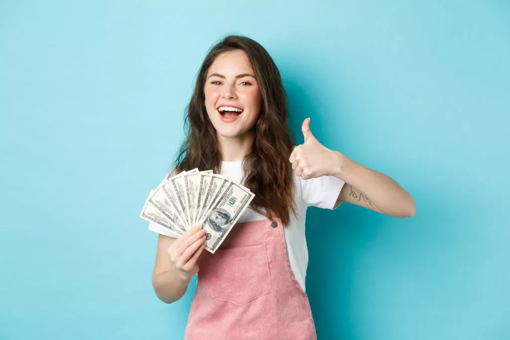 Smiling happy woman holding money, dollar bills and showing thumb up, recommending fast cash loan and looking satisfied, standing over blue background